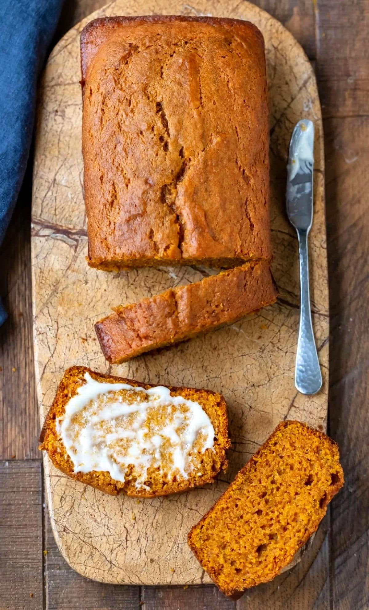 A loaf of freshly baked pumpkin bread on a wooden cutting board, with one slice buttered and two other slices beside it. A butter knife rests next to the bread, highlighting the moist texture and golden-brown crust