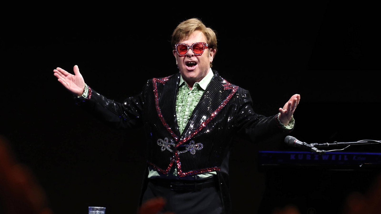 Elton John performing on stage, wearing his signature colorful outfit featuring a black sequined jacket with red accents and heart-shaped sunglasses. His arms are spread wide as he sings into a microphone on a Kurzweil keyboard. Known for his iconic style and legendary music career, Elton captivates audiences worldwide with his charismatic presence. This image highlights his vibrant energy and unique flair that have made him a global superstar in the music industry.